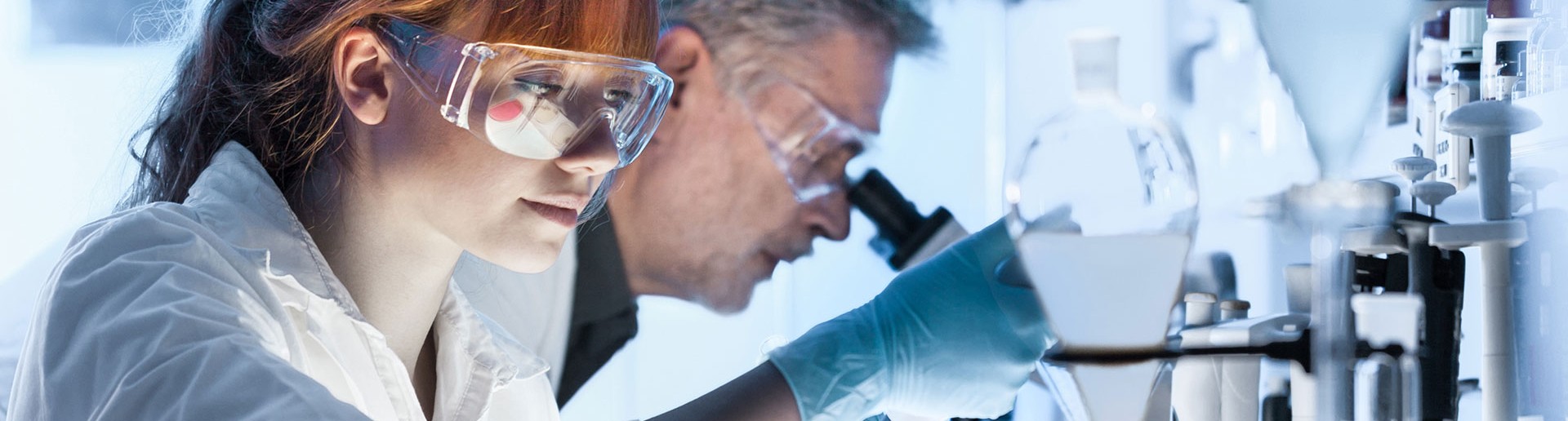 Químico de laboratorio sonriente con gafas y, en el fondo, un microscopio y tubos de ensayo.